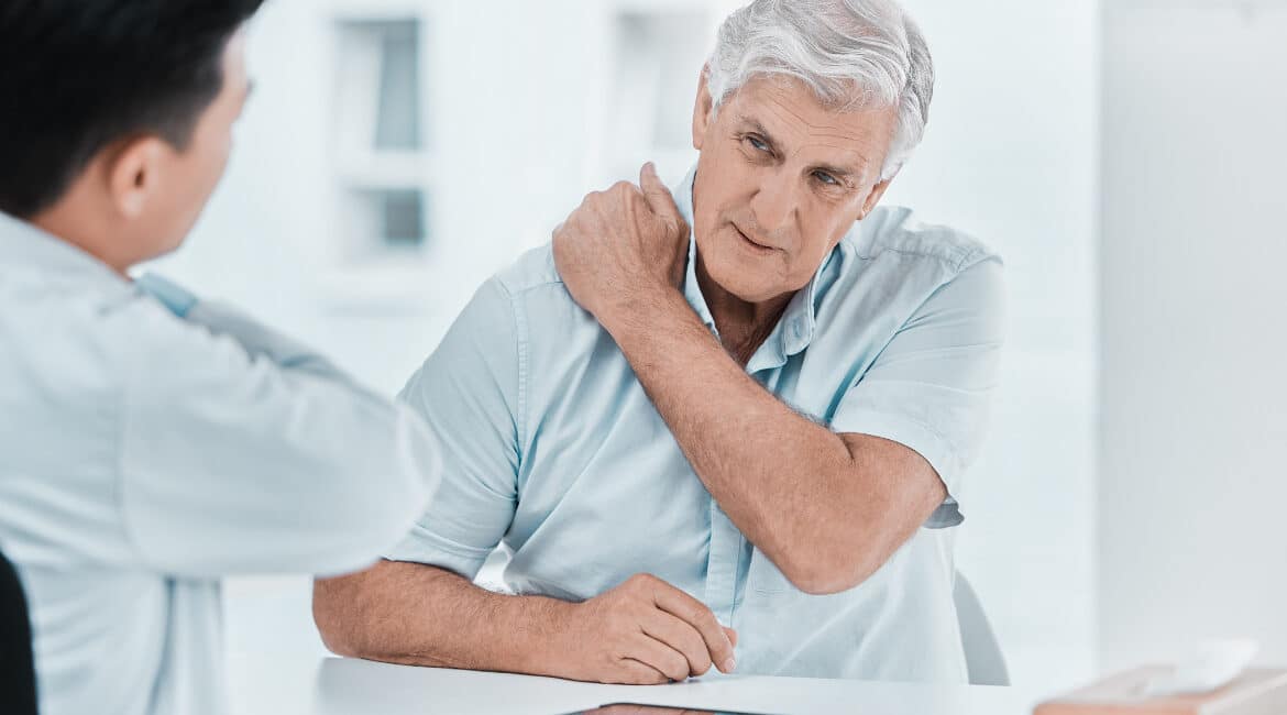 An elderly man in a light blue shirt talks to a doctor about his shoulder pain in a bright medical office. Revision orthopedic surgery is discussed.