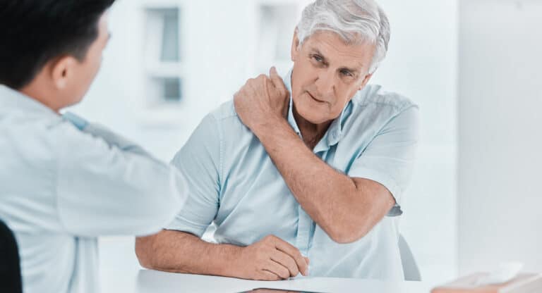 An elderly man in a light blue shirt talks to a doctor about his shoulder pain in a bright medical office. Revision orthopedic surgery is discussed.